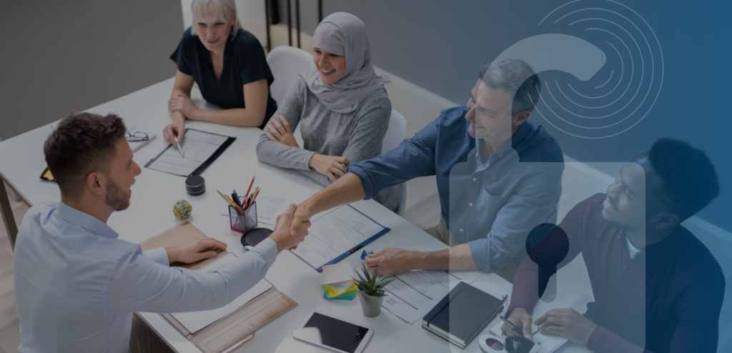 A diverse group of business professionals gathered around a meeting table, engaged in a collaborative discussion. One man shakes hands with another across the table.