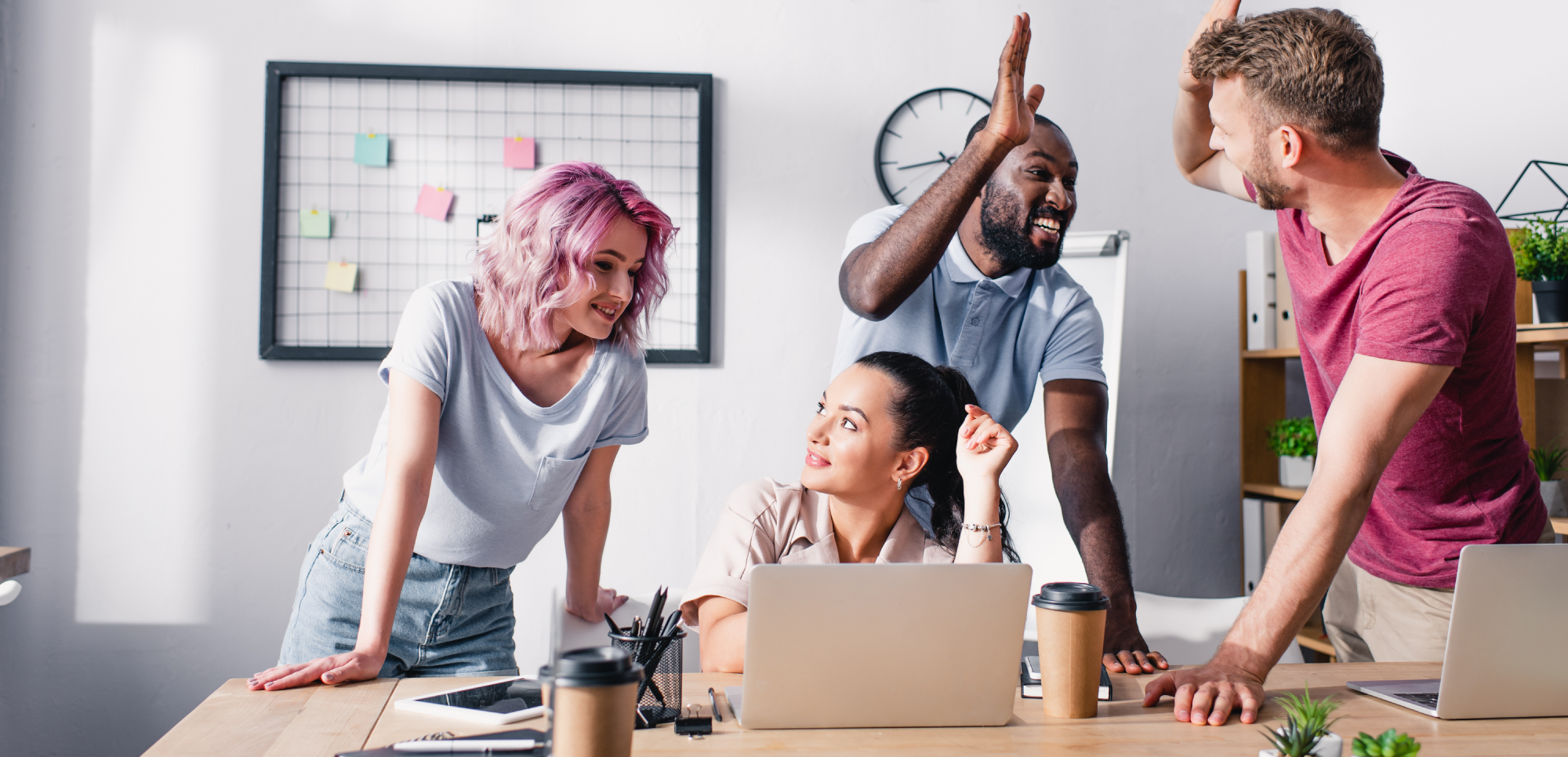 A diverse team of four, including a woman with pink hair and a man giving a high-five, collaborating around a modern office desk. They appear engaged and happy, surrounded by laptops and personal notes, in a bright workspace with a wall-mounted idea board in the background.