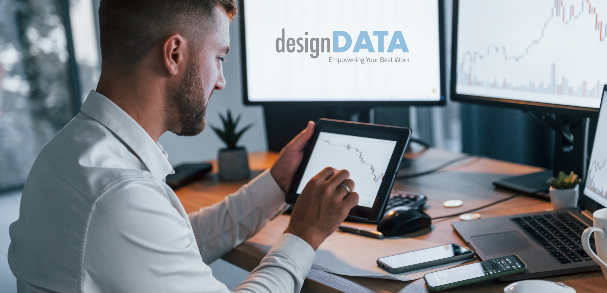 A professional man analyzes data on a tablet, sitting at a desk surrounded by multiple screens displaying graphs.