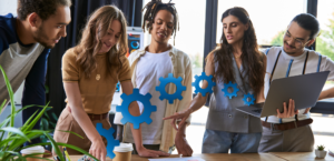 A vibrant team of five young professionals, diverse in appearance and style, engaged in a discussion over a workbench. They point at documents and a laptop, surrounded by graphic icons of gears, symbolizing workflow and innovation. The setting is a bright, modern office with large windows.