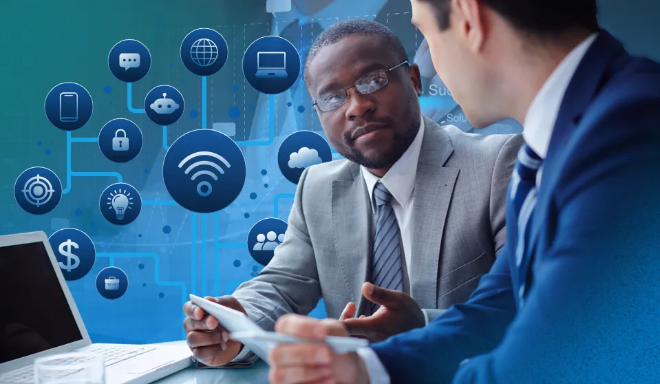 An image that represents the title of the blog: How Much Should Nonprofits and Associations Spend on Technology? There are two men sitting in front of a laptop in business clothes. Behind them, various icons representing their IT infrastructure are floating in the background: WiFi network symbol, cloud, etc.